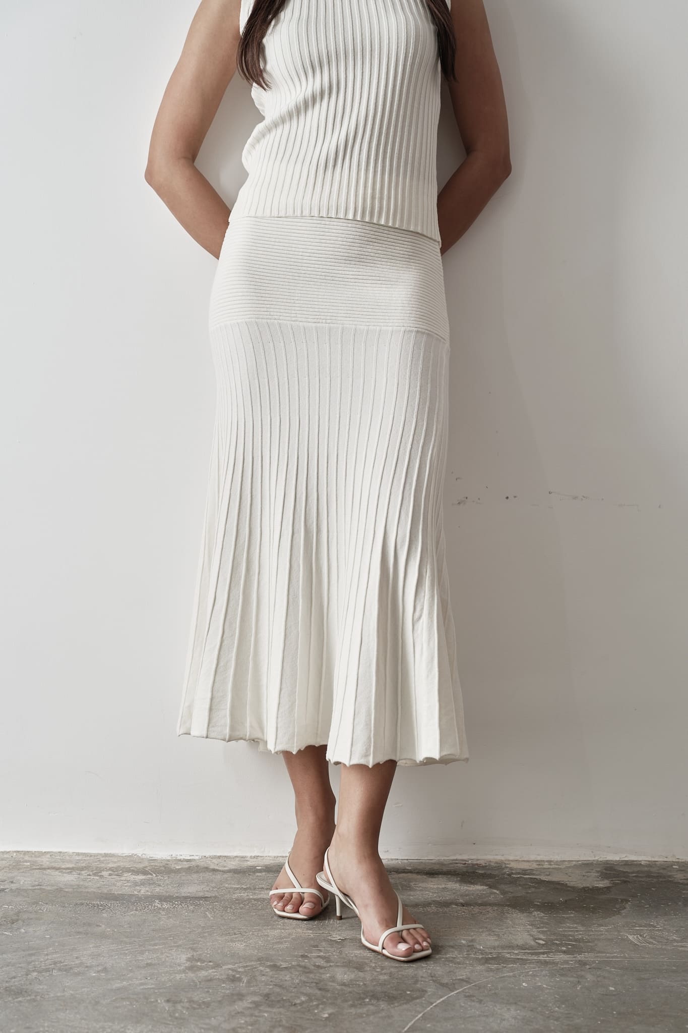 Model wearing Andrea Set, featuring a ribbed sleeveless top and a matching pleated midi skirt, styled with white strappy sandals, posing against a white background