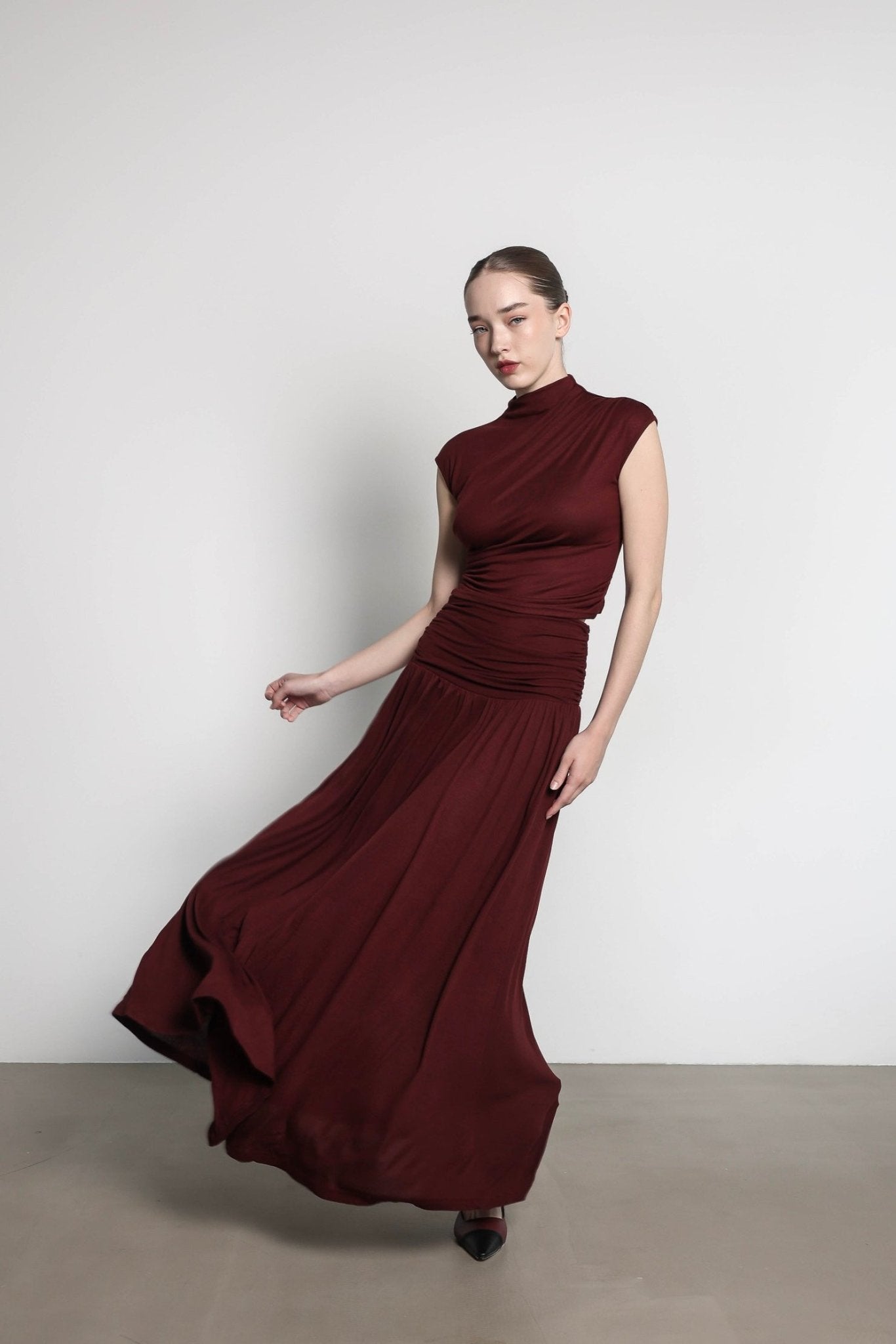 A woman in the Aria set, featuring a flowing burgundy maxi skirt, poses against a plain white backdrop. She stands on a neutral-colored floor, her skirt swirling elegantly around her legs. Wearing black shoes and with her hair pulled back, the overall tone is minimalist and sophisticated.