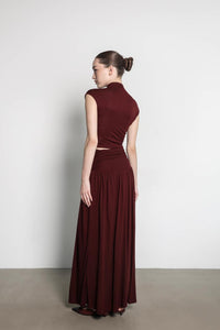 A woman with dark brown hair in a bun is standing against a plain white background. She is wearing the Aria set, featuring a sleeveless dark red top and a matching long, flowing maxi skirt. She is facing slightly to the side with her gaze to the right.