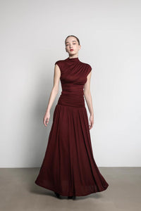 A person stands against a plain white background, wearing the Aria set, which includes a high-necked top and a floor-length burgundy skirt. The form-fitting top complements the loosely flowing maxi skirt. The individual has their arms relaxed by their sides.