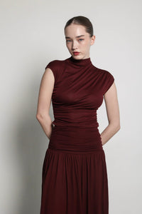 A woman with fair skin and straight brown hair pulled back stands against a plain background. She wears a fitted, sleeveless burgundy high-necked top from the Aria set. Her hands are clasped behind her back, and she gazes forward with a neutral expression.