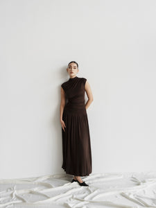 A person stands against a plain white wall on draped white fabric, wearing the Aria set, which includes a sleeveless dark brown floor-length maxi skirt with subtle draping and black shoes. Their hair is pulled back, and they have a neutral expression while slightly leaning on the wall.