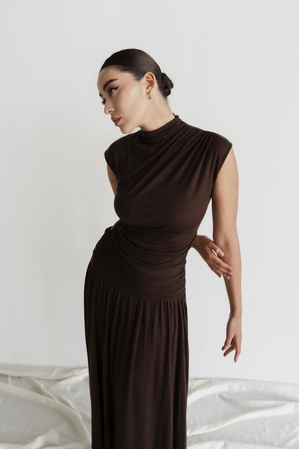 A person with slicked-back hair stands in a side pose against a plain background, showcasing the Aria set—a fitted dark brown dress with cap sleeves. One arm is folded behind their back while the other rests relaxed at their side, striking a graceful pose.