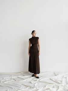 A woman stands against a white background, wearing a fitted, sleeveless dark brown dress from the Aria set that falls to her ankles. The elegant maxi skirt style accentuates her figure as she has her hair pulled back and sports black shoes. The floor beneath her is draped in white fabric. With one arm relaxed by her side, she looks to her left.