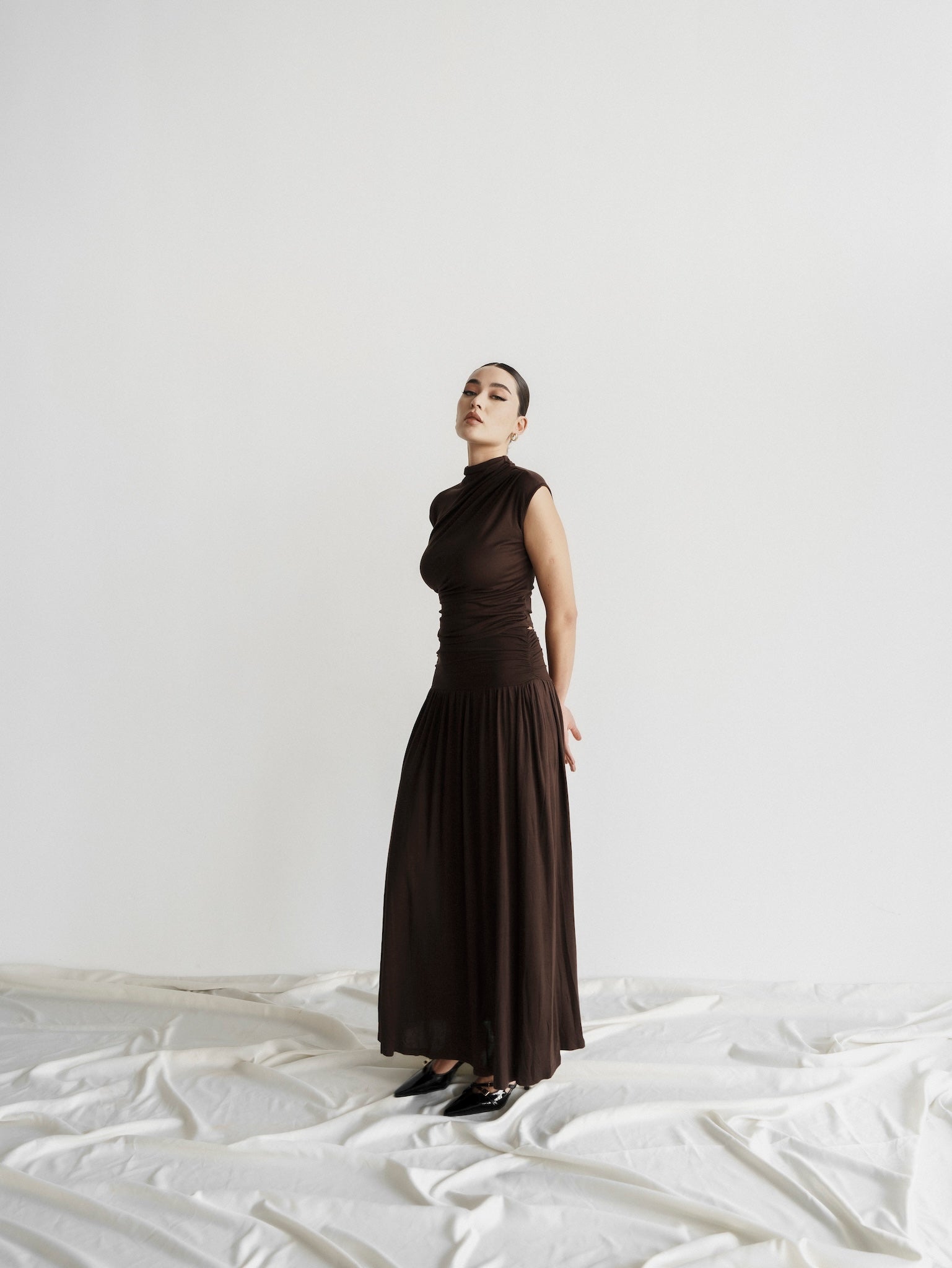 A woman stands against a plain white background, dressed in the elegant Aria set—a dark brown, sleeveless, high-necked top that flows seamlessly into a floor-length gown with a slightly ruched bodice. She has her hands behind her back and looks directly at the camera. The floor is covered with white fabric.