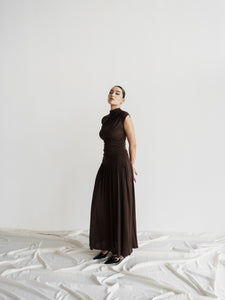 A woman stands against a plain white background, dressed in the elegant Aria set—a dark brown, sleeveless, high-necked top that flows seamlessly into a floor-length gown with a slightly ruched bodice. She has her hands behind her back and looks directly at the camera. The floor is covered with white fabric.