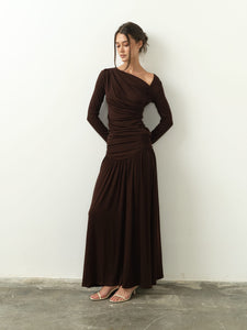 A woman stands against a plain white wall, wearing the Ariana Set—a long-sleeved, asymmetrical dark brown dress featuring a draped design and an asymmetrical neckline. She has her hands behind her back and complements the outfit with light-colored sandals. Her hair is styled in a loose bun.