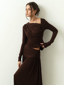 A woman wearing the refined Ariana Set, featuring a long-sleeved brown dress with an asymmetrical neckline, stands against a plain backdrop. Her right hand rests on her hip as she gazes to the side with a serene expression. Her hair is styled in a loose updo, enhancing her elegant appearance.