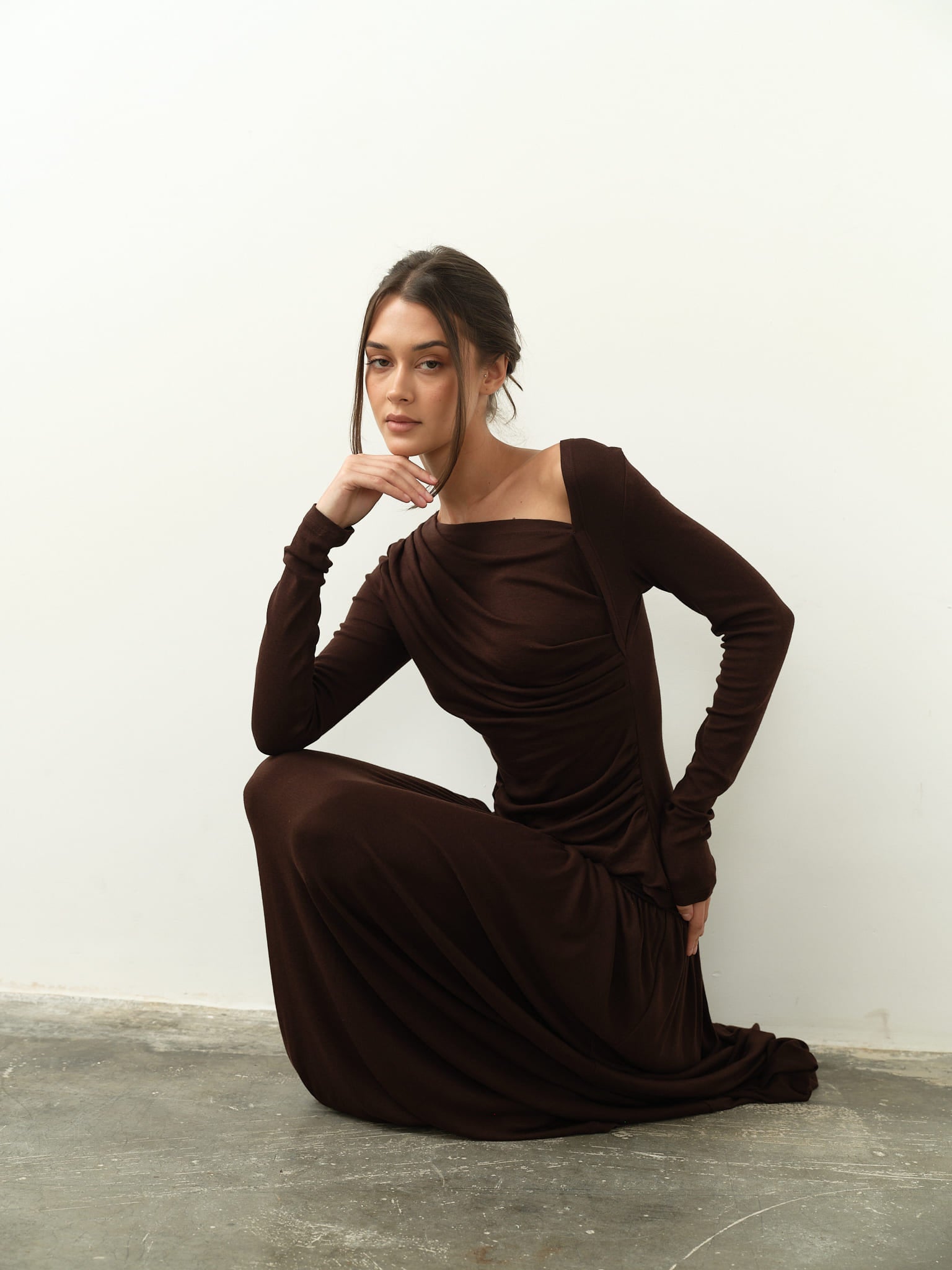 Dressed in a dark brown Ariana Set, a woman poses gracefully against a plain white wall, kneeling with one hand on her knee and the other gently touching her chin. Her thoughtful expression is accentuated by the full-length skirt, which adds an elegant contrast to the plain gray concrete floor.