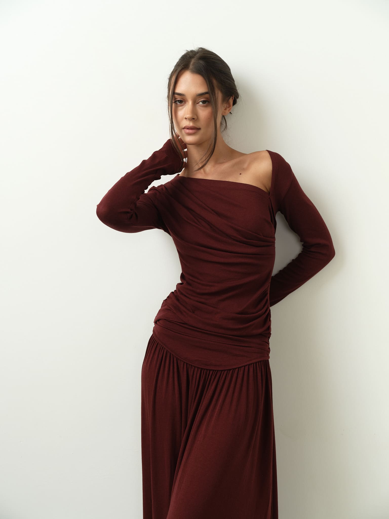 Wearing the Ariana Set, a woman stands against a white wall in a long-sleeved, off-shoulder outfit with an asymmetrical neckline. Her right hand gently touches her cheek, her hair is tied back, and she displays a relaxed expression.