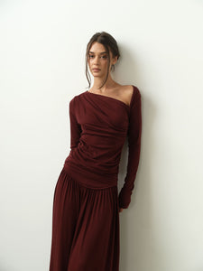 A person with long hair is dressed in the Ariana Set, characterized by a burgundy color, an asymmetrical neckline, and matching wide-leg pants, as they lean against a plain white wall. The soft lighting enhances the sophisticated and elegant ambiance.