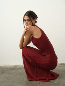 A person crouches on a concrete floor, dressed in an Asteri Dress featuring a sleeveless, halter neckline. They gaze thoughtfully at the camera with one hand resting on their chin. The background is a plain off-white wall.