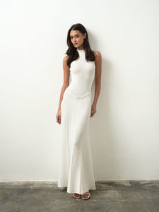 A woman wearing the chic, floor-length Asteri Dress poses against a simple white background. Her long, dark hair cascades as she gazes sideways, highlighting the dress's form-fitting silhouette and halter neckline for an elegant and minimalist appearance.