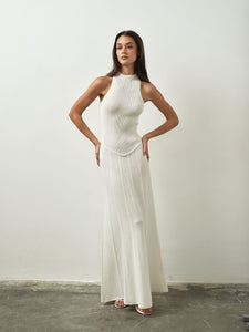 In a minimalist room, a woman poses confidently against a plain white wall, wearing the Asteri Dress—a sleeveless, long white maxi dress featuring a halter neckline. Her long hair complements the form-fitting top of the dress, which elegantly flares slightly towards the bottom as she stands with her hands on her hips.