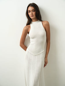 A woman with long, dark hair poses against a white wall, donning the sleeveless, high-neck Asteri Dress. Her serene expression enhances the elegance of the maxi dress's silhouette as her left arm bends gracefully behind her back.