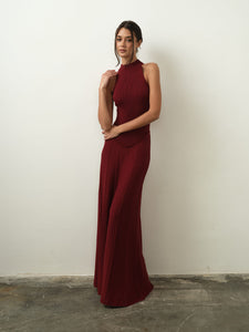 An individual wearing a sleeveless Asteri Dress in deep red stands before a plain white wall. The dress boasts a high halter neckline and a long, flowing skirt. Their long dark hair is styled in a casual updo.