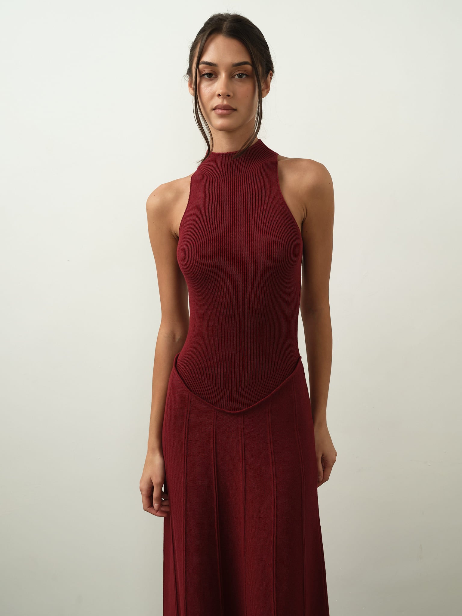 A woman poses against a simple white backdrop, wearing the Asteri Dress in sleeveless, deep red. The dress boasts a halter neckline and transitions into a long skirt, capturing the graceful essence of a maxi dress. Her naturally styled long, dark hair completes the look.