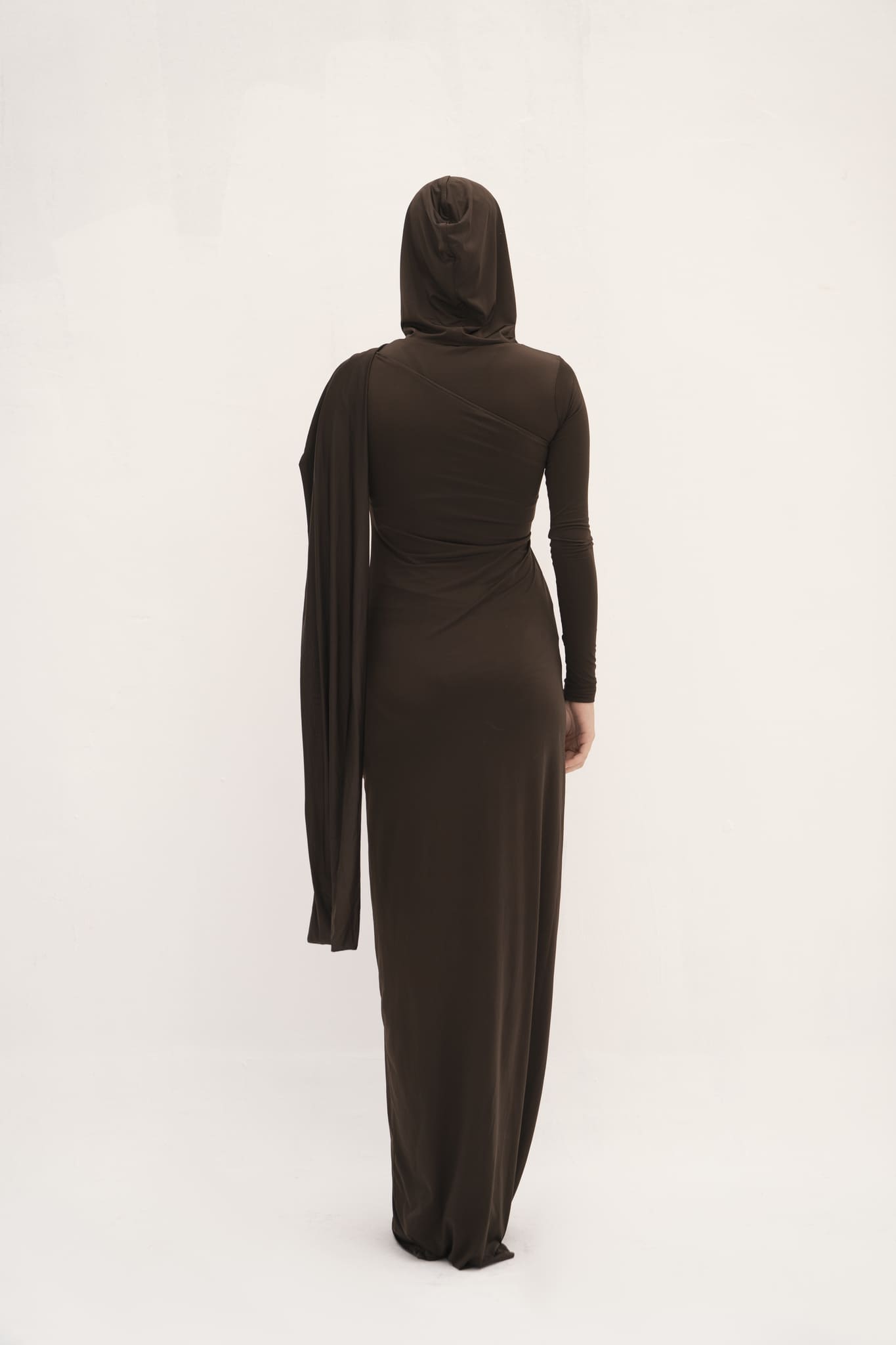 This dress has a sleek, fitted design in a dark, earthy tone with long sleeves and an attached drape that can be wrapped around the head, creating a sophisticated and modest look. The fabric appears smooth, with a slightly matte finish, and the overall silhouette is elongated and elegant. 
