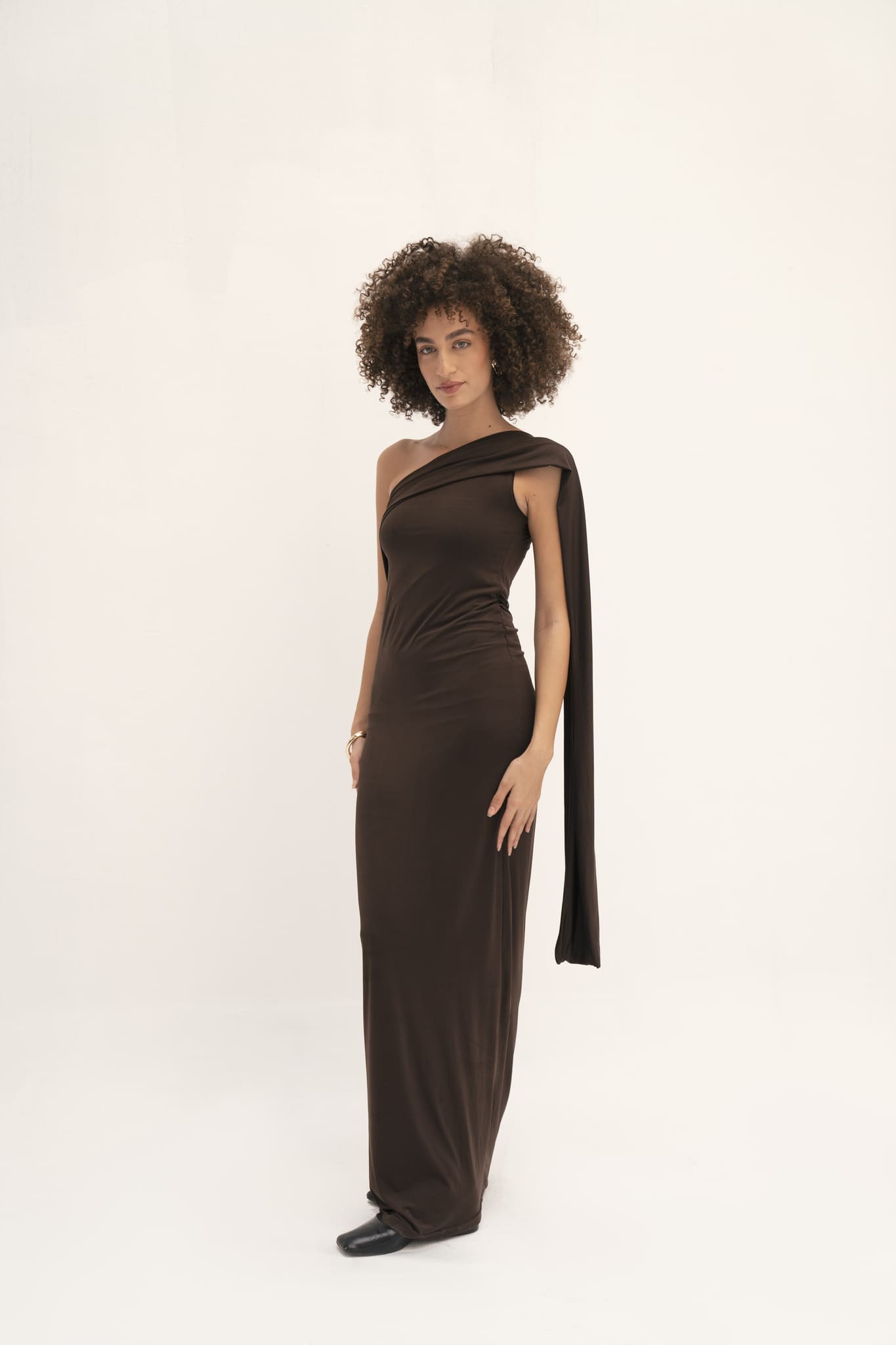 A person with curly hair is standing against a plain white background, wearing the Ava Gardner Dress—a sophisticated, form-fitting must-have in dark brown with long draped shoulder details. They complete their look with black shoes.