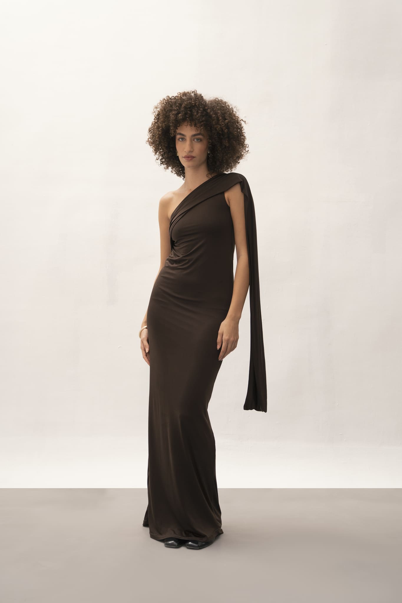 A person with curly hair is elegantly posed against a simple light background, wearing the Ava Gardner Dress—a sophisticated essential. This long, dark one-shoulder dress is highlighted by its draped embellishment, making it an ideal and versatile choice for any refined event.