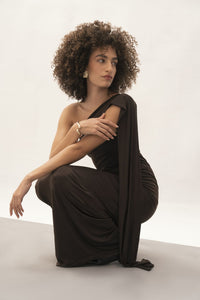 A person with curly hair poses elegantly in the chic must-have Ava Gardner Dress, sitting on a stool. Against a plain light backdrop, they cross their arms adorned with statement earrings and a bracelet.