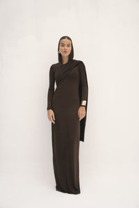 A woman stands against a plain white background, elegantly dressed in the Ava Gardner Dress—a dark brown, long-sleeved, floor-length piece paired with a head covering and a gold bracelet. This sophisticated ensemble perfectly complements her composed expression as she gazes directly ahead.