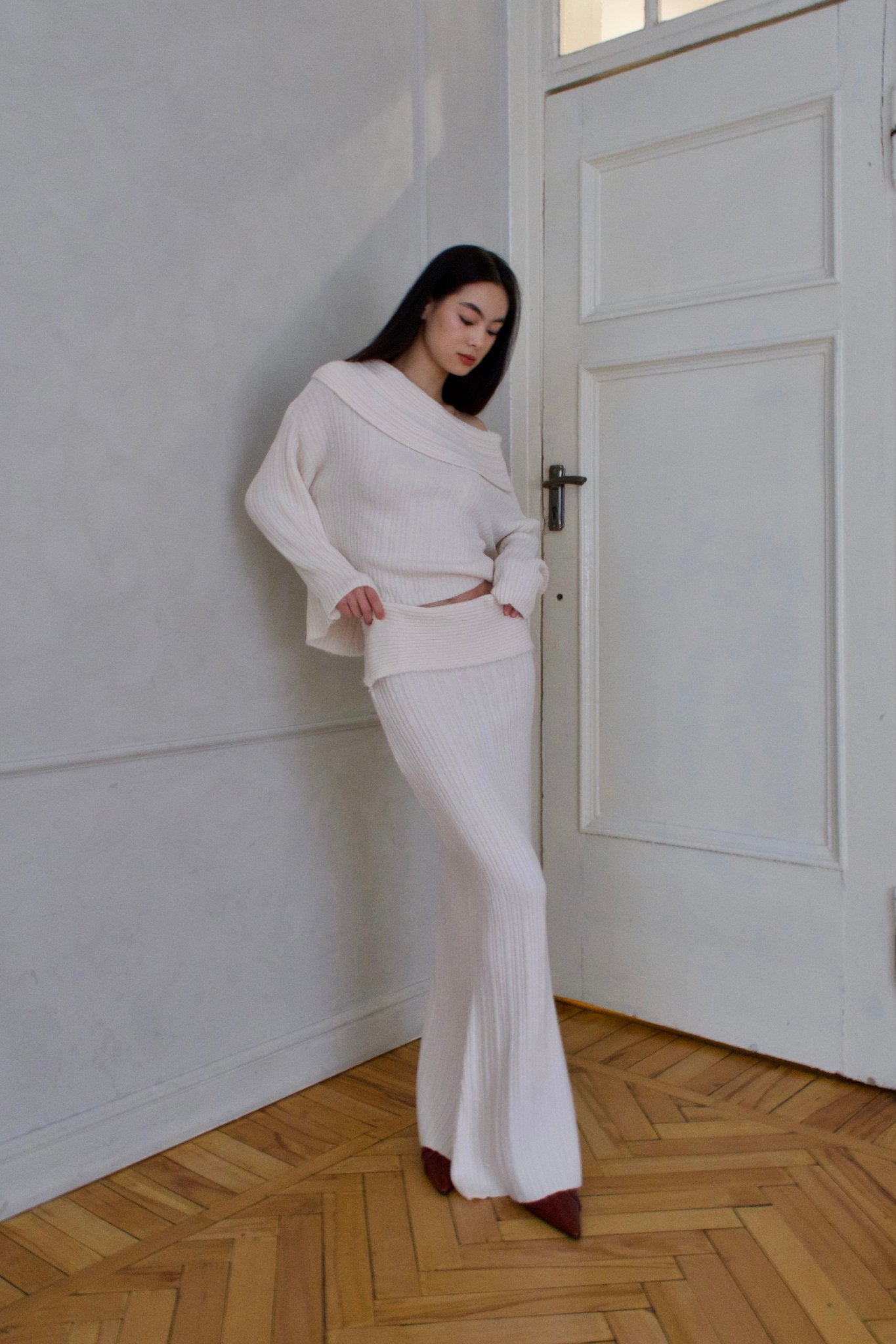 A woman with long dark hair is standing indoors, leaning against a white door with her hands resting on her hips. She is wearing the Ayla set, which includes an off-the-shoulder, long-sleeve knitwear top and matching high-waisted ribbed pants. She has paired this outfit with pointed brown shoes.