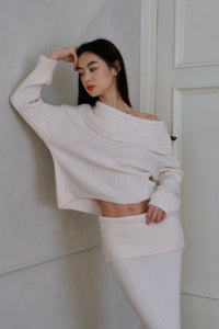 A woman with long dark hair leans against a wall in an Ayla set, consisting of an off-the-shoulder, oversized cream-colored sweater paired with a matching ribbed skirt. With one arm raised and her hand resting on her forehead, she looks pensive. This chic knitwear outfit is available for international delivery.