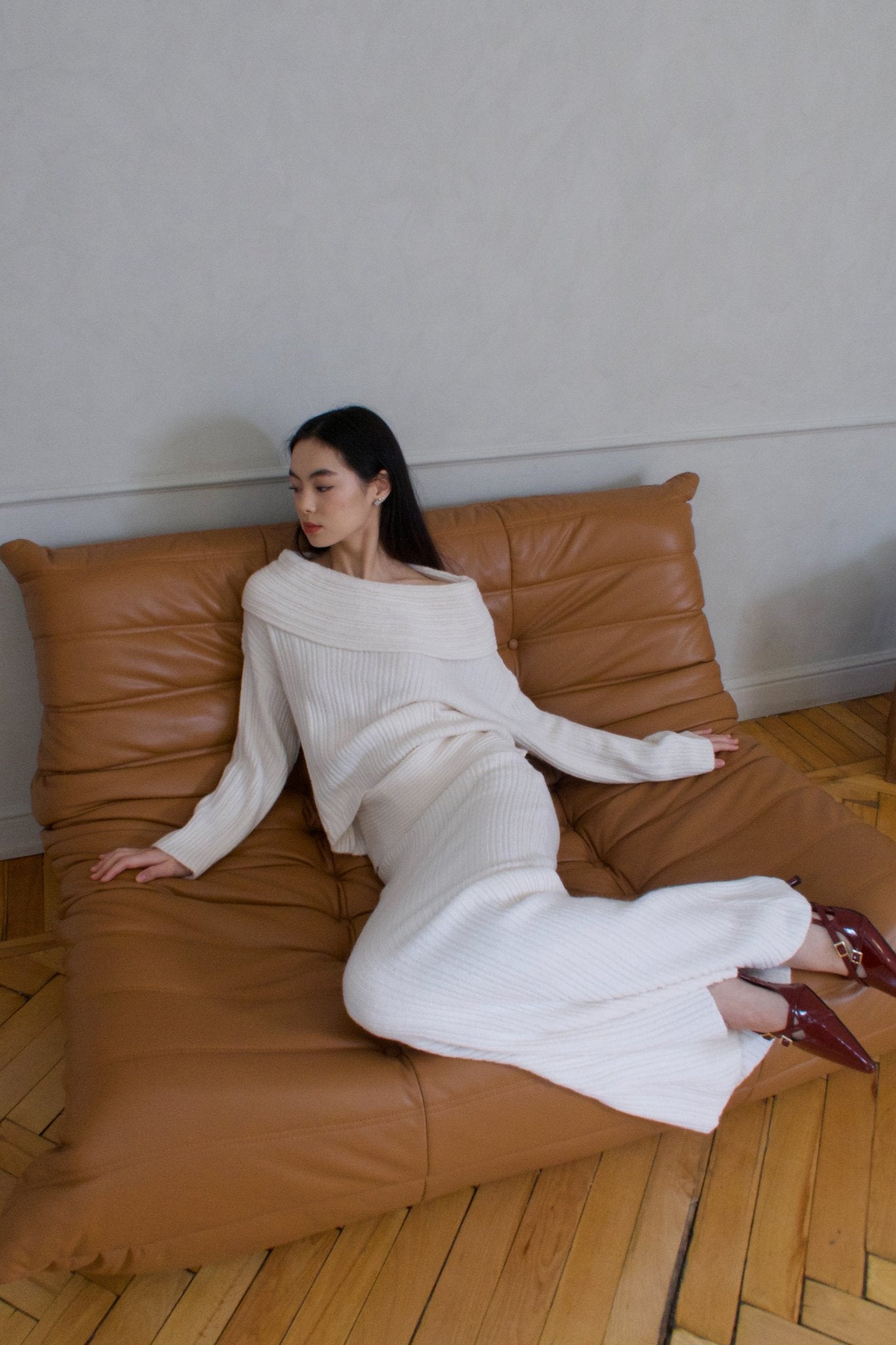 A woman dressed in an Ayla set of white knitwear reclines on a minimalist tan couch positioned on a wooden floor, with a light-colored wall as the backdrop. She gazes to the side, her hands resting on the seats.