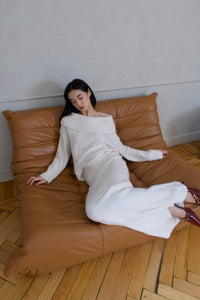 A woman dressed in the Ayla set, consisting of a white ribbed off-shoulder sweater and matching long skirt, sits on a brown leather couch with a relaxed posture. She leans slightly to one side, with her dark hair cascading loosely over her shoulders. This knitwear ensemble made from breathable fabric complements the wooden floors beautifully.