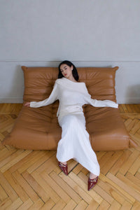 A woman with long dark hair is lounging on a tan leather couch. She is dressed in a cozy, off-the-shoulder white knitwear piece from the **Ayla set** and a matching white knit skirt. She is wearing pointed burgundy shoes with gold details. The background features light-colored walls and herringbone wooden flooring.