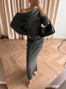 A person dressed in the Ayla set—a stylish ribbed gray off-the-shoulder sweater and matching skirt—is taking a mirror selfie. They have a black handbag slung over their shoulder, standing on a wooden floor with beige curtains in the background. This chic knitwear ensemble is available for international delivery.