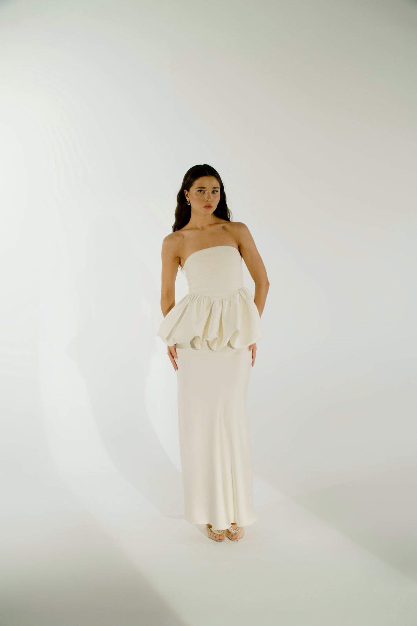 A woman with long, dark hair stands against a plain background wearing the Bella Top in white. This strapless silhouette features a ruffled waist detail. She accentuates her look with earrings and high-heeled shoes while looking slightly to the side.