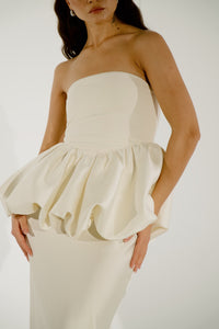 A woman is seen from the shoulders down, wearing an elegant, off-white strapless silhouette. The dress features a fitted bodice and a dramatic ruffled waist from the Bella Top, flowing into a slim-fitted skirt. Her hands rest gently on the ruffle.