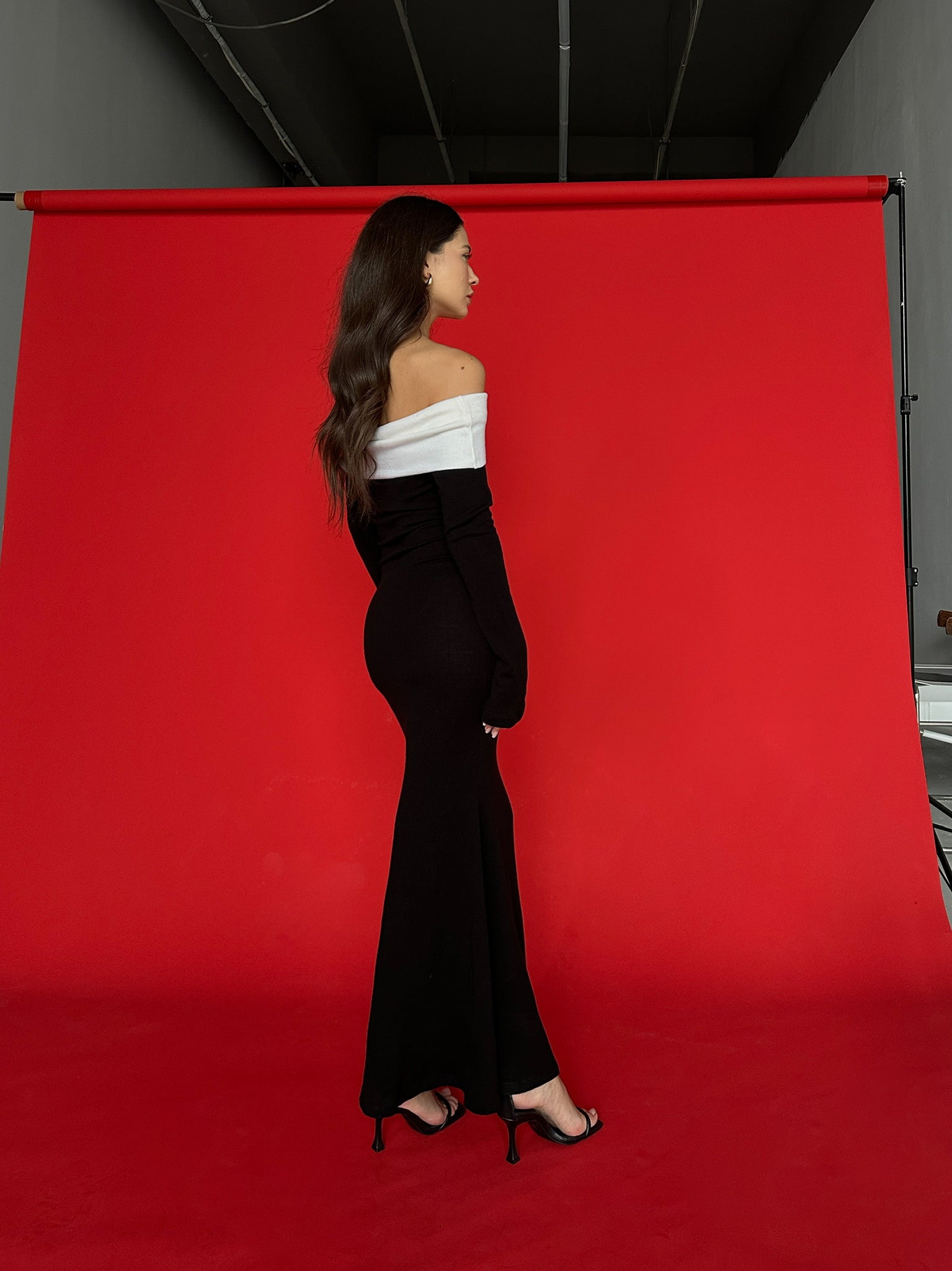 A woman with long, dark hair stands in profile against a red backdrop. She is wearing the Esber knit dress, a body-hugging black off-the-shoulder design with a white band across the top, paired with black high-heeled sandals. The setting appears to be an indoor studio.