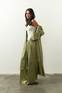 Draped in the sophisticated Talisa Set, a woman presents an elegant look with a silky satin robe and wide-legged pants set against a neutral backdrop. Her long dark hair pairs beautifully with the white sleeveless top under the flowing green robe as she stands gracefully on a concrete floor, exuding refined charm.