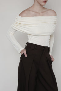 A close-up shot of a young woman wearing a cream-colored off-the-shoulder top with ruched detailing. The top elegantly drapes across her shoulders and arms, highlighting the soft texture of the fabric. She pairs it with high-waisted, wide-leg brown trousers, which feature pleats and a structured waistband. Her hands are casually tucked into the pockets of the trousers, adding a relaxed yet sophisticated feel to the outfit. The neutral tones and minimalist style emphasize a refined, modern look.







