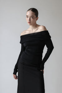 A young woman is wearing a sleek, black off-the-shoulder long-sleeved top paired with a long black skirt. Her hair is neatly styled in a tight bun, slightly serious expression. The minimalist and elegant outfit highlights her sophisticated and modern fashion sense.