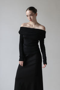 A young woman is wearing a sleek, black off-the-shoulder long-sleeved top paired with a long black skirt. Her hair is neatly styled in a tight bun, slightly serious expression. The minimalist and elegant outfit highlights her sophisticated and modern fashion sense.