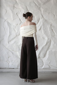 A young woman is photographed from behind, showcasing her off-the-shoulder cream-colored top with ruched detailing along the back and sleeves. She pairs the top with high-waisted, wide-leg brown trousers, which elongate her silhouette. Her hair is styled in a smooth, elegant bun. She completes the outfit with white strappy heels. The textured white wall in the background adds a modern and minimalist touch to the overall aesthetic, highlighting the clean lines of the outfit.







