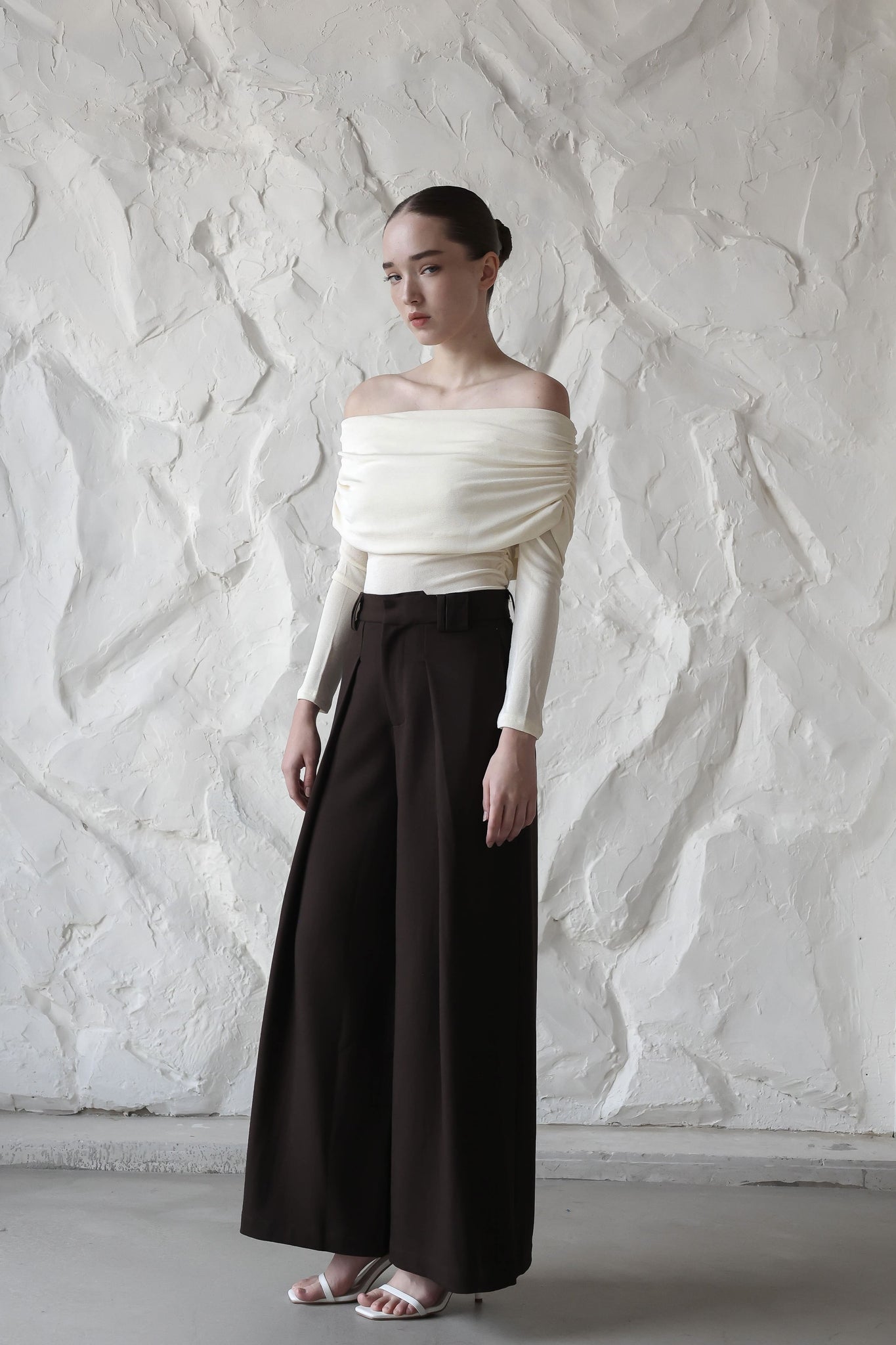 A woman with her hair tied back is standing against a textured white wall. She is wearing the sophisticated Camélia top, high-waisted wide-leg black pants, and white sandals. She is looking slightly to the side with a neutral expression.