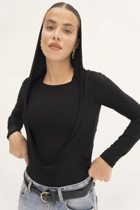 In front of a neutral backdrop, an individual wears the Carolyn Top in black, featuring a draped hood over their shoulders, paired with light blue jeans. This jersey knit fabric top beautifully complements their hoop earrings and confident demeanor, as they place their hands on their hips.