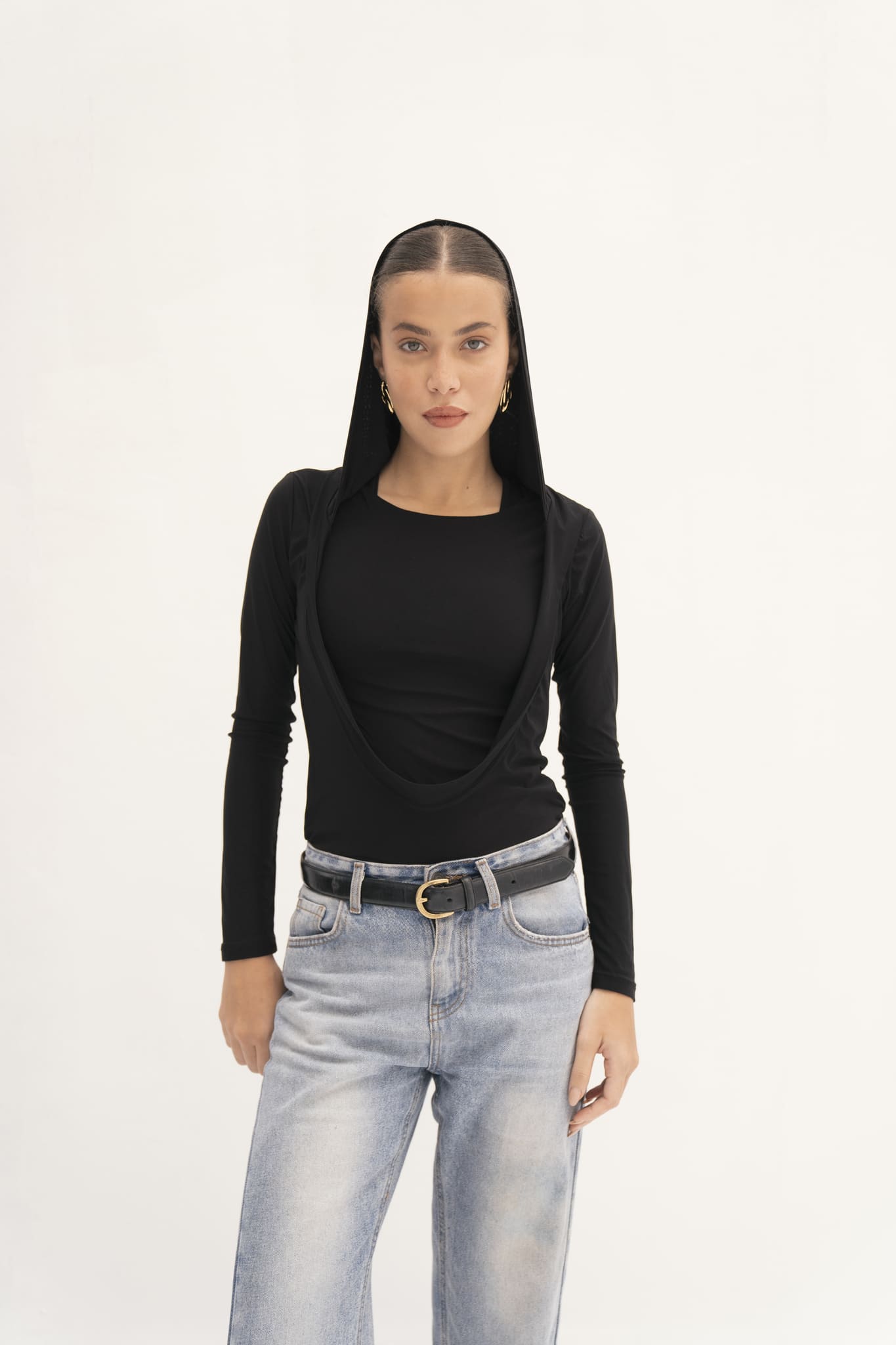 A person wearing a Carolyn Top in black, featuring a draped hood, paired with light blue jeans is set against a plain white background. The ensemble, crafted from soft jersey knit fabric, is accessorized with a belt and hoop earrings. Their expression remains neutral.