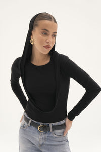 A person models the Carolyn Top, featuring a draped hood over their head, accessorized with large gold earrings and paired with light blue jeans secured by a black belt. The jersey knit fabric enhances their confident expression as they pose against a plain white background.