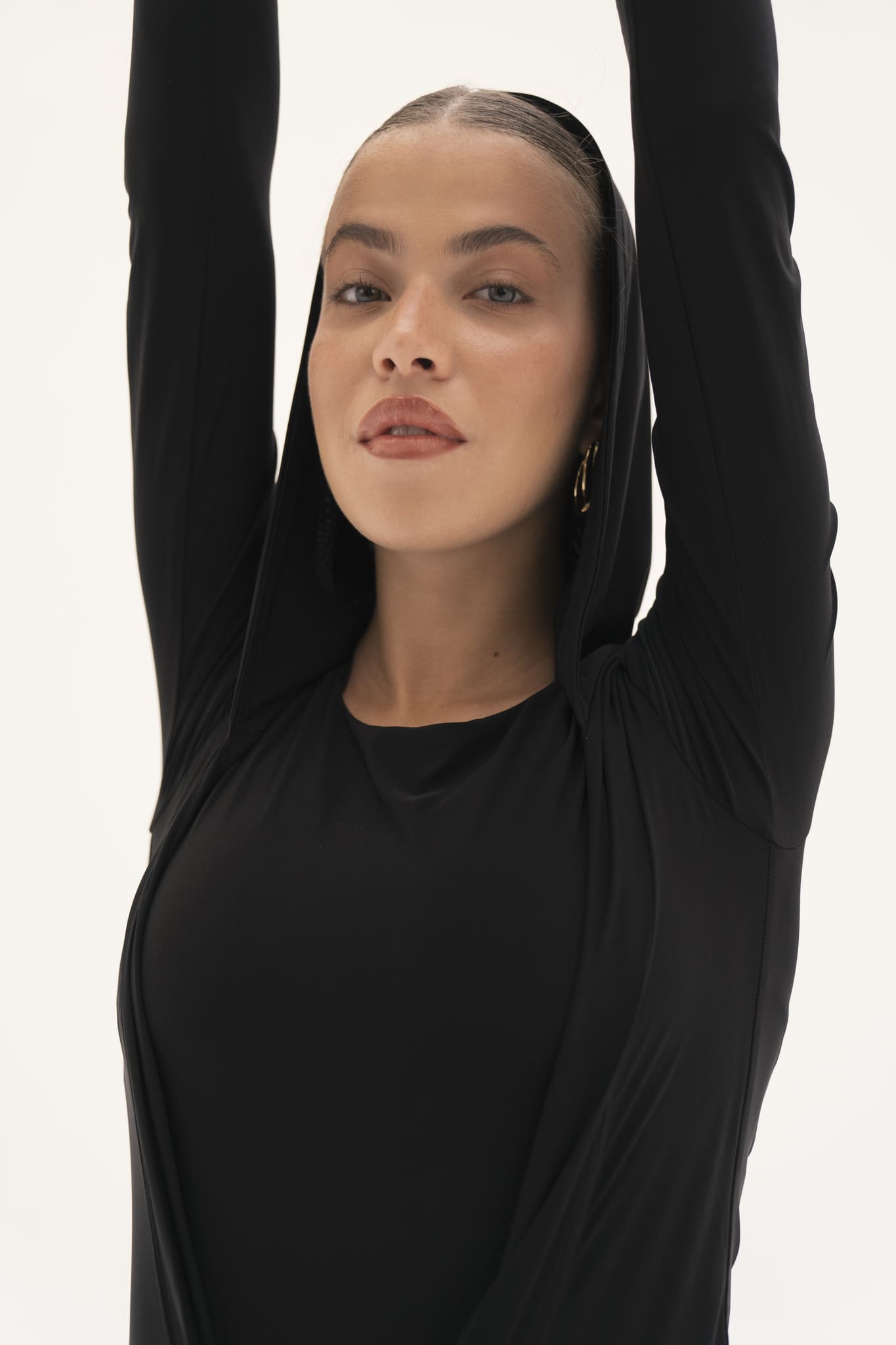 A person wearing the Carolyn Top in black jersey knit fabric stretches with arms raised above their head, looking directly at the camera against a plain background.