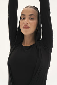 A person wearing the Carolyn Top in black jersey knit fabric stretches with arms raised above their head, looking directly at the camera against a plain background.