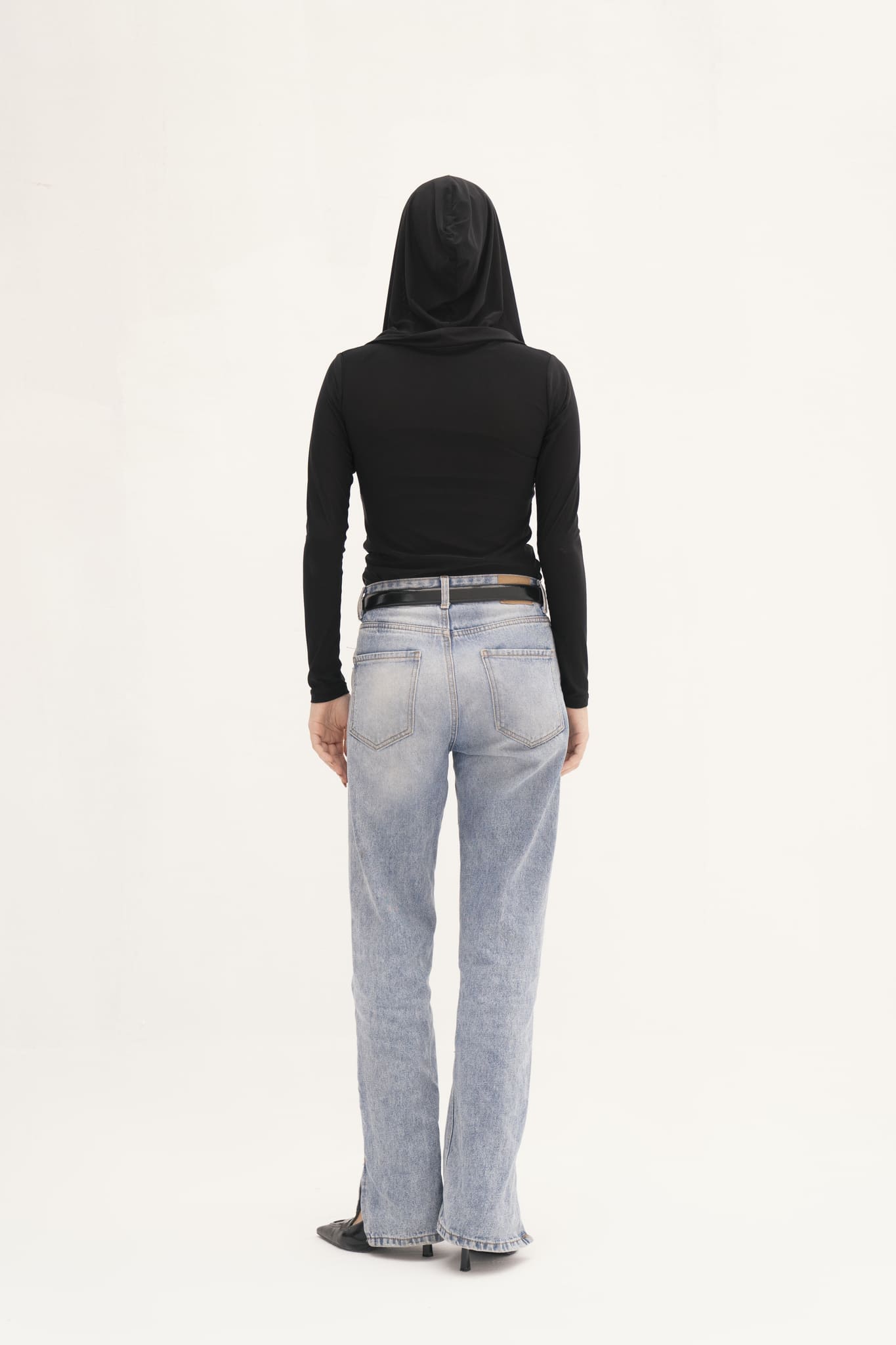 A person wearing a Carolyn Top in black, featuring a draped hood, paired with blue jeans and black shoes, stands facing a plain white background.