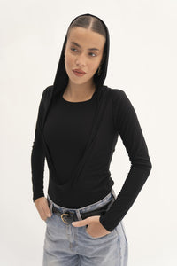 A person stands confidently with their hands in their pockets, wearing a Carolyn Top in black, featuring a draped hood, paired with light blue jeans. The jersey knit fabric enhances both comfort and style. A plain, light-colored background accentuates the sleek appeal of the outfit.