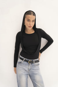 A person with long brown hair wears the Carolyn Top in black jersey knit, showcasing a draped hood over their head. Paired with blue jeans and a black belt, they stand against a plain white background, gazing thoughtfully to the side.