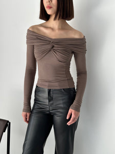 A person stands against a plain white background, wearing the Sabella top with long sleeves and a twisted front detail, crafted from high-quality soft cotton fabric. Pairing it with black leather pants, their straight shoulder-length hair frames their face as their hands are relaxed by their sides.